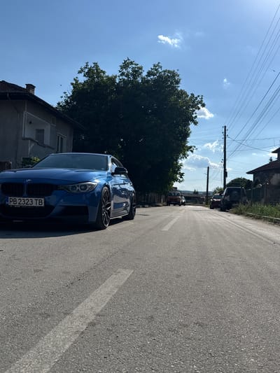 A blue BMW sedan is parked on the left side of a paved road in Strazhitsa, Bulgaria. The car's license plate reads "PB 2323 TB". To the left of the car is a grey residential building with a tiled roof and a chimney. A large green tree is positioned behind the car. The road extends into the distance, where several other vehicles are parked, including an orange van and a dark-colored SUV. Power lines are visible stretching across the sky. The sky is blue with scattered white clouds, and the sun is shining. The scene appears to be a quiet residential street on a sunny day.