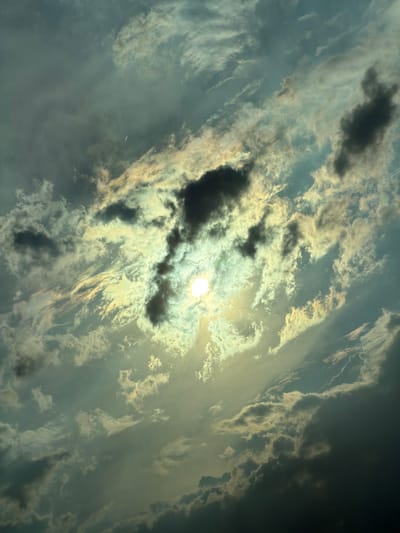 The image depicts a dramatic sky filled with a mix of dark storm clouds and bright, sunlit clouds. The sun is partially obscured by clouds, creating a luminous halo effect and casting rays of light through the atmosphere. The clouds exhibit a range of textures, from wispy cirrus formations to denser cumulus clouds, some appearing dark and brooding, while others are illuminated with golden and white hues. The overall impression is one of dynamic weather, possibly transitioning between stormy conditions and sunshine. The perspective is as if looking upwards, with the clouds filling the entire frame. The image captures a moment of intense natural beauty and atmospheric drama. No people, objects, or text are visible in the image. The time of day is suggested to be either sunrise or sunset due to the dramatic lighting and cloud colors. Given the context of Gavrailovo, Bulgaria, this scene likely represents a natural sky formation over a rural or semi-rural landscape. The emotions evoked by the image could range from awe and wonder to a sense of foreboding due to the presence of dark clouds.