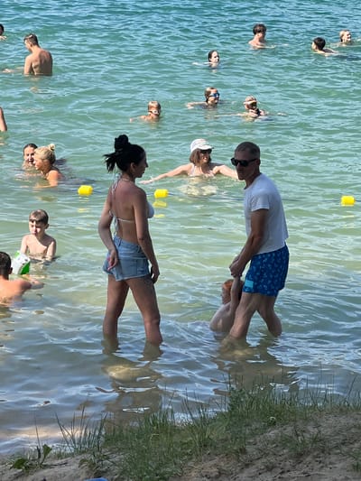 This image captures a sunny day at a lake, with several people of various ages swimming and interacting in the shallow water. The water is a clear, light turquoise, and the seabed is visible in places, suggesting a sandy or gravelly bottom. The shoreline in the foreground is sandy, with sparse green grass growing.
In the mid-ground, a woman in a light-colored bikini and denim shorts stands facing left, with her back to the camera. To her right, a man in a white t-shirt and blue patterned swim shorts is holding the hand of a young child, who appears to be red-haired. The man is facing the woman. To the left of the woman, a young boy in swimming trunks is visible, looking down at a green object in the water. Further back, several other individuals are swimming and enjoying the water. Some are further out in the lake, while others are closer to the shore. Yellow buoys are present in the water, possibly marking a swimming area.
While the image depicts a pleasant scene at a lake, it does not provide any specific visual cues that would allow for identification of the location as being within the city of Vilnius, Lithuania. The environment appears to be a generic lake or large body of water suitable for recreation.