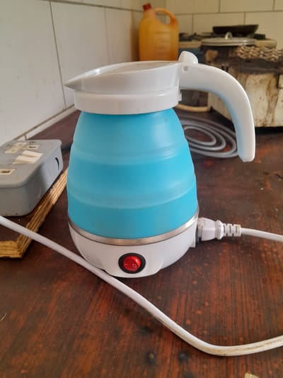 A blue collapsible electric kettle with a white lid and handle sits on a dark wooden table. The kettle is plugged into a power outlet. To the left of the kettle is a grey electrical outlet box. In the background, there is a yellow jug and what appears to be a stove. The scene is indoors.