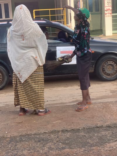 In Shagamu, Nigeria, a person wearing a patterned headscarf and a long, patterned skirt stands to the left. Their back is mostly to the camera, with a portion of their patterned skirt visible. To the right, another person, dressed in a dark, floral-patterned long-sleeved shirt and dark trousers, holds a broom and a shallow basket containing food items. This individual is wearing an orange-brimmed green cap and orange flip-flops. They are facing away from the first person and are looking upwards, with their right arm raised. They are standing next to a dark-colored car, with its rear wheel visible. The ground is a dirt and gravel surface. In the background, a building with windows and a yellow metal railing is visible. A red and white sign is also present on the building.