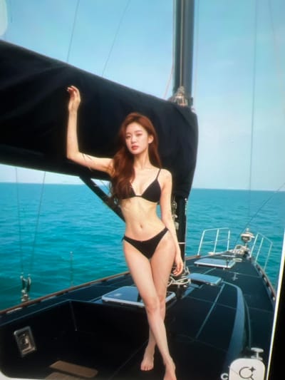 The image shows a woman wearing a black bikini standing on a sailboat. She has long, brown hair and is looking at the camera. Her left arm is raised, resting on a black sail behind her. The sailboat is on a body of water with a clear blue sky above. The water appears to be calm and turquoise. The deck of the sailboat is black and has metal railings. The image is taken from a slightly low angle, emphasizing the woman and the sailboat against the backdrop of the sky and sea. The location in the city of Kara-Dıykan, Kyrgyzstan is not evident from the image.