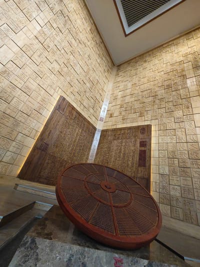 A low-angle, wide shot captures an indoor scene in the Daxing District of China. Walls are covered in beige square tiles, each etched with various symbols, presumably Chinese characters. Two large, dark wooden panels also bear inscriptions. In the foreground sits a large, round, brown object with radial divisions, resting on a stone surface. The room is well-lit, with overhead lighting and subtle spotlights highlighting the walls. The overall setting suggests a cultural or historical exhibition within a museum or similar institution. No people are visible.