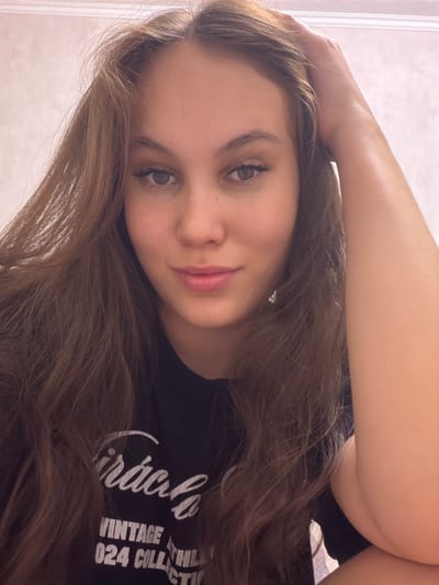 A young woman with long, brown hair is captured in a close-up, head-and-shoulders shot. She is looking towards the camera with a slight smile. Her right hand is raised and resting on top of her head, with her fingers extended. She is wearing a black t-shirt with white text on it, which is partially obscured by her hair and arm. The background is a plain, light-colored wall with a subtle texture. The lighting suggests an indoor setting. No specific location in Naberezhnye Chelny, Russia can be determined from the image.