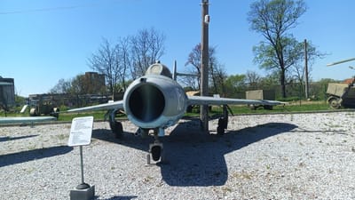 The image captures a static display of a MiG-17 fighter jet in Yambol, Bulgaria, set in an outdoor area covered with gravel. The aircraft, painted in gray, dominates the foreground. The cockpit is visible, and the jet has a large, circular intake. An identification or information sign is positioned on the left side of the jet, mounted on a pole. Various pieces of military equipment are scattered in the background. Bare trees and trees with green foliage are present in the background, along with utility poles. The sky is blue, suggesting a clear day.
