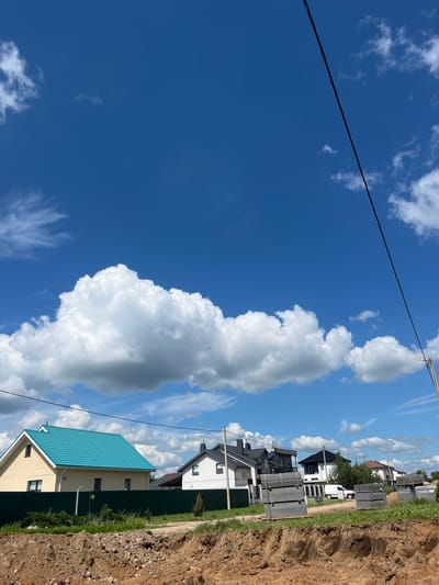 The image depicts a residential area under development in the Бараўлянскі сельскі Савет, Belarus. In the foreground, a raw, excavated dirt area with loose soil and rocks suggests ongoing construction. Beyond this, a dark green metal fence encloses a section of properties.
Several houses are visible. The leftmost house has a light yellow facade and a striking bright turquoise roof. Next to it, a white house with a dark roof is situated. Further to the right, a cluster of houses with dark roofs and modern architectural styles are present, one of which has a white facade. A white van is parked in front of some of these houses. In front of some of the houses, stacks of grey construction materials, possibly concrete blocks or slabs, are organized. The ground behind the fence appears to be grass and vegetation.
The sky is a vibrant blue with large, fluffy white cumulus clouds and some wispy cirrus clouds. A power line stretches diagonally across the upper right portion of the frame, connected to a pole visible in the far right. The overall scene suggests a sunny day in a developing suburban or rural environment.
