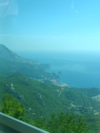 An aerial, wide-angle, slightly hazy view captures the coastal landscape of Brajici, Montenegro. The scene is dominated by lush green, densely forested mountains that cascade down to meet the bright blue Adriatic Sea. A serpentine road winds its way through the hills, with scattered buildings and structures visible amidst the trees and along the coastline. The sea itself is calm, with a few small boats dotting the surface and a distant, hazy horizon line where the sea meets the pale blue sky. The image appears to have been taken from a moving vehicle, possibly a bus or car, due to the blurred foliage in the foreground and the reflection of what looks like a window frame in the upper left corner.