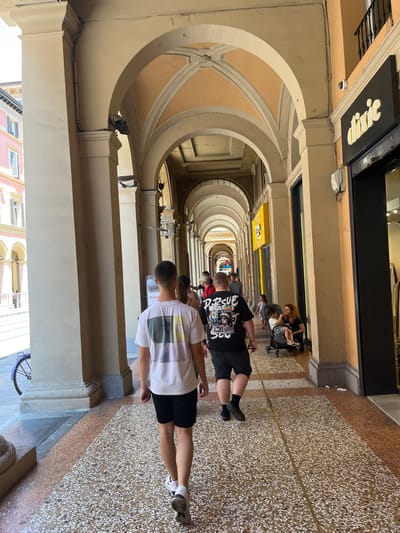 The image shows a long, covered walkway in Bologna, Italy, characterized by repeating arches that create a tunnel-like perspective. The walkway is paved with a speckled pattern of white and brown stones. A few people are walking along the walkway. Two young men are walking in the foreground. One is wearing a white t-shirt and black shorts; the other is wearing a black t-shirt and black shorts. Further down the walkway, a person pushes a stroller. Stores line one side of the walkway with signs visible. One sign reads "Dixie." A security camera is visible above one of the arches. The walkway is lit by natural light filtering through the arches and openings. The image is taken at ground level, looking down the length of the walkway.