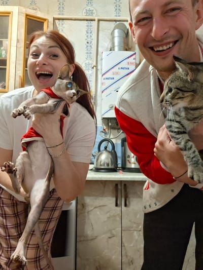 A man and a woman are holding cats in what appears to be a kitchen. The woman, on the left, has red hair and is wearing a white t-shirt and plaid pants. She is holding a hairless cat, possibly a Sphynx, wearing a red garment. The man, on the right, is smiling and wearing a light-colored jacket with red accents. He is holding a tabby cat. In the background, there is a white gas water heater with blue and red lettering, a kettle, and kitchen cabinets. The setting seems to be domestic, possibly a home in the village of Забалоцкі сельскі Савет, Belarus, given the presence of the water heater with Cyrillic text.