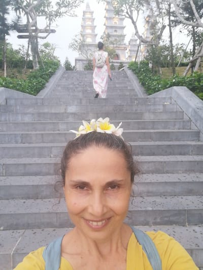 A selfie shows a middle-aged woman with a crown of frangipani flowers in her hair standing at the bottom of a long, wide, stone staircase leading to a religious structure in the background. The woman has dark hair and light brown skin. She is wearing a yellow shirt with a light blue strap over her shoulder. She is smiling at the camera. The staircase extends upwards and appears to lead towards a temple-like structure with multiple tiered pagodas. The steps are grey and show signs of being damp or wet. In the background, a woman in a pink and white patterned dress and a backpack is ascending the stairs. The stairs are lined with greenery. Trees frame the sky above. The photograph was taken in Hong Gai Ward, Vietnam.