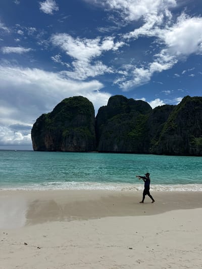 A lone individual stands on a white sand beach in Ao Nang, Thailand, pointing to the left. The person wears a dark blue long-sleeved shirt, black pants, and a dark baseball cap. They are barefoot. The turquoise water of the ocean gently laps the shore, with small waves visible. In the background, towering, lush green limestone karst islands rise dramatically from the sea under a bright blue sky dotted with white, fluffy clouds. The scene captures a moment of serene natural beauty characteristic of the region.