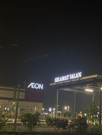 The image, captured at night in Bekasi Regency, Indonesia, depicts a brightly lit commercial establishment. A prominent, illuminated sign displaying the text "SELAMAT JALAN" (meaning "Goodbye" or "Farewell" in Malay/Indonesian) is visible towards the top-right. Directly beneath it, a vertical illuminated sign features the "AEON" logo and name, suggesting a shopping mall or large retail complex.

These signs are mounted on an overhead structure, possibly an entryway canopy or part of the building's facade. In the foreground, a dark metal mesh fence runs across the lower portion of the frame, separating the viewer from the scene. Behind this fence, various plants and shrubs with green and red foliage are visible, subtly illuminated by the ambient light. Further in the background, multiple streetlights or facility lights emit a soft glow against the dark night sky.

The left side of the image is predominantly dark, but on this dark surface, faint, inverted, and somewhat ghostly reflections of both the "SELAMAT JALAN" and "AEON" signs are discernable, indicating a reflective surface nearby or a lens effect. The overall scene is devoid of people, conveying a static, nighttime view of a modern commercial area.