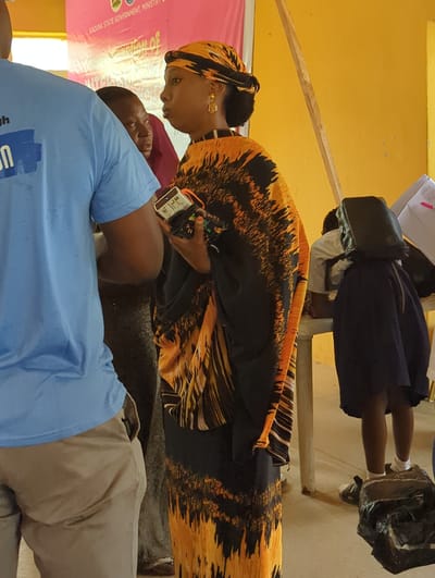 In Kakuri Gwari, Nigeria, a woman in an orange and black patterned dress and headscarf stands holding a mobile device. To her left, a person wearing a blue t-shirt and khaki pants is partially visible. In the background, a woman in a purple headscarf and another person wearing a white shirt are seen. A young girl in a dark blue skirt and white socks stands to the right. The environment appears to be indoors, with a yellow-painted wall and a table visible. The scene suggests a gathering or event.