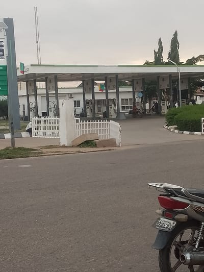 The image shows a gas station in Pantame, Nigeria. A white motorcycle is parked in the foreground, with its license plate visible. In the background, a series of gas pumps are lined up under a covered structure. The sky is overcast, and the scene appears to be outdoors. There are a few people visible in the background, some of whom appear to be employees of the gas station. The overall impression is one of a typical day at a gas station in Nigeria.