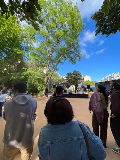 The image captures a scene in Lisbon, Portugal, likely in a park or public square. People are gathered, some standing and facing forward, as if observing a performance or event taking place on a stage in the distance. There is a stage with people on it in the center of the image. Buildings are visible in the background. Trees with green leaves are scattered throughout the scene, some directly overhead. The sky is blue with white clouds.