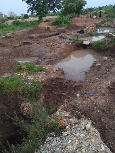 The image depicts a landscape in Otulu, Nigeria, characterized by a disturbed terrain suggestive of excavation or mining operations. Several pools of murky water are present, interspersed with mounds of excavated earth and debris. Patches of green vegetation, including grass and trees, are visible, contrasting with the predominantly brown and reddish hues of the soil.
In the mid-ground, three individuals are present. One person, wearing dark clothing, stands near a pool of water. Another individual, also in dark attire, appears to be working near a pile of sand. A third person, dressed in yellow and white, is also engaged in activity in the vicinity. Various objects, including blue hoses, what appear to be pipes, and possibly equipment for pumping or sifting materials, are scattered throughout the scene. The overall impression is one of an active, though rudimentary, industrial or agricultural site. The sky is overcast, suggesting potentially subdued lighting conditions.