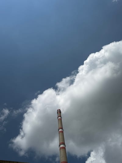 An upward-angled shot captures a tall industrial chimney against a partly cloudy sky in Pirdop, Bulgaria. The chimney, a prominent object in the upper right portion of the frame, is constructed from concrete or similar material, featuring distinct red and light grey or white bands near its top section. It extends diagonally across the sky, suggesting its immense height.

The sky is a mix of deep blue and large, voluminous clouds. A significant mass of greyish-white clouds occupies the upper and right-hand side of the image, appearing thick and somewhat turbulent. These clouds partially obscure the chimney, and what appears to be an emission (likely steam or smoke) from the chimney's top blends into the cloud cover, creating an atmospheric merging of industrial output with natural weather phenomena. The lower left of the frame showcases a clearer, vibrant blue sky, contrasting with the heavier cloud cover. The overall impression is of a bright daytime, with diffuse lighting indicating either an overcast sky or bright conditions diffused by the clouds. No people are visible, and no discernible text appears on the chimney or in the sky. The scene highlights the scale of industrial infrastructure set against a vast sky.