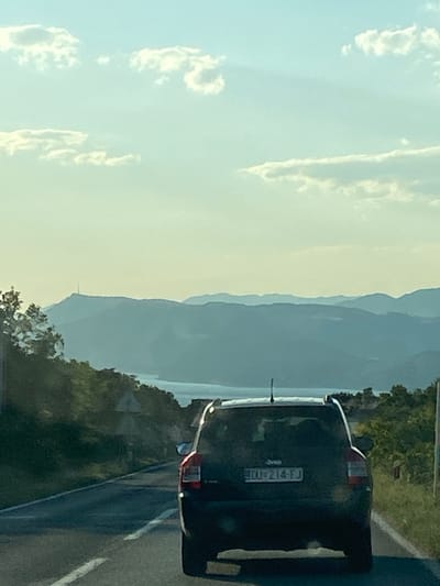 The image shows the rear of a dark-colored SUV traveling on a paved road in Močići, Croatia. The license plate number DU-214-FJ is visible on the vehicle. A line of trees and vegetation borders the road on the right. In the background, mountains are visible under a bright, slightly cloudy sky.
