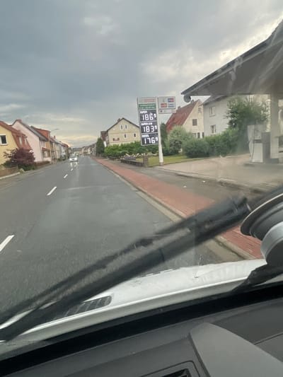 The image shows a street scene in Wartenberg, Germany. The view is from inside a vehicle, looking forward through the windshield and wipers. On the right side of the image is a gas station with a price sign displaying fuel costs. The sign indicates "SUPER 1.86", "SUPER E10 1.83", and "DIESEL 1.76". Houses line the street on both sides. The sky is cloudy and appears overcast. A white car is visible driving down the road in the distance. The road is wet, suggesting recent rain.
