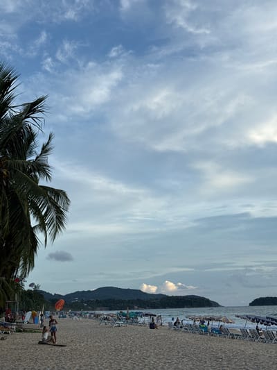 This image depicts a crowded beach scene in Karon, Thailand. The scene is dominated by a sandy beach lined with rows of lounge chairs and umbrellas, suggesting a popular tourist destination. In the foreground, the fronds of palm trees are visible against a sky filled with scattered clouds. The ocean is a shimmering expanse, with gentle waves lapping at the shore.

Several people are scattered across the beach, some lounging on chairs, others walking or engaged in activities. In the middle ground, two individuals are visible, one on a surfboard and another standing beside them. A colorful parachute, likely for parasailing, is also present, hinting at recreational activities available. Further down the beach, more people and beach paraphernalia are visible, creating a sense of a lively atmosphere.

The sky suggests it is either late afternoon or early evening, with a soft, diffused light. The weather appears to be pleasant, with a mix of clouds and clear sky. In the background, lush green hills rise from behind the beach, providing a scenic backdrop. The overall impression is one of a tropical vacation spot, bustling with activity and enjoying the coastal environment. There is no visible text in the image.