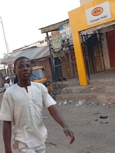 The image depicts an unpaved commercial street in Gama, Nigeria, under diffused daylight. In the foreground, a young male, dark-skinned, wearing a light-colored V-neck tunic and trousers, walks towards the camera, looking directly forward. His right arm is visible, positioned as if in motion. Behind him, a partially visible yellow three-wheeled vehicle is present. To the left, a second male subject, dressed in white with a white cap, walks away from the camera.
The background consists of a row of single-story commercial buildings, many featuring corrugated metal roofs. A prominent yellow structure on the right identifies as an "MTN Connect Point" with its logo and signage, accessible via concrete steps. Various advertisements and signs are affixed to the buildings; one partially legible blue sign reads "GAMA", and another advertises "ALL NETWORK AIRTIME". An indistinct dark shape lies on the ground near the buildings in the mid-ground right. The ground surface is dirt or gravel and is littered with plastic bags and other refuse. A tall, slender utility pole stands on the far left.