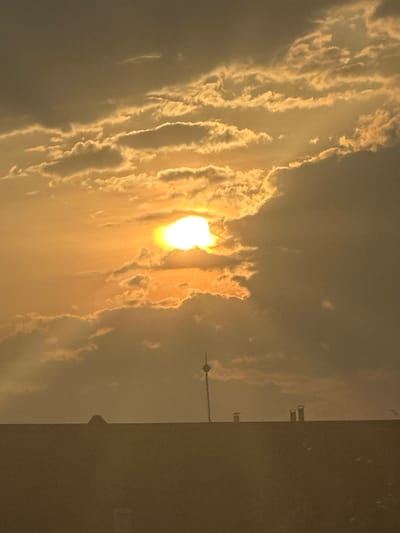 The image, oriented vertically, captures a dramatic sky during either sunrise or sunset over Graf Ignatievo, Bulgaria. The central focus is a brilliant, radiant sun, partially obscured by dynamic clouds, casting an intense golden-yellow light across the scene. These clouds, which fill the majority of the frame, exhibit intricate patterns and textures, with their edges glowing brightly where the sun hits them directly, while other sections appear darker and shadowed.
On the right side of the image, against the illuminated horizon, several structures are visible in silhouette. A prominent feature is a tall, slender communication tower or mast, resembling those found at an air base, topped with a distinctive spherical or disc-like component. Further to the right, additional industrial-looking buildings or structures, possibly with chimneys, are also silhouetted against the vivid sky. The very bottom portion of the image is a dark, undefined mass, likely representing the ground or a building rooftop, providing a stark contrast to the luminous sky above. The scene conveys a sense of serene natural beauty and the captivating interplay of light and shadow during a pivotal part of the day. No text is visible in the image.
