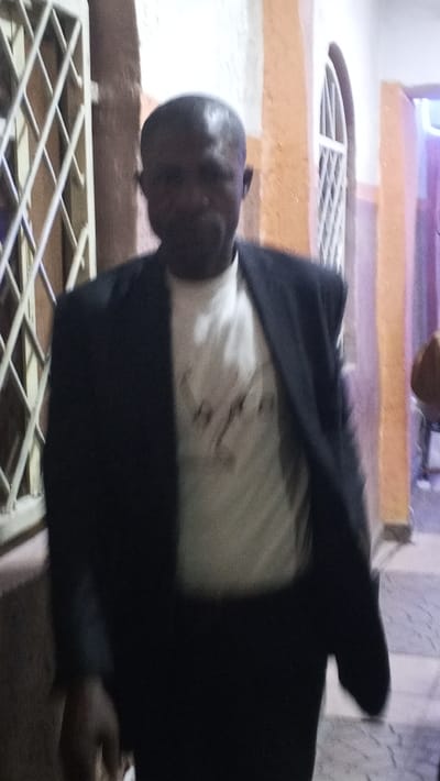 The image depicts an adult male in Sabon Gari East, Nigeria, captured in mid-stride within an indoor or sheltered environment. The subject, appearing to be middle-aged, possesses dark skin and a receding hairline. He is dressed in a dark blazer over a light-colored t-shirt featuring an indiscernible dark graphic design on the chest, paired with dark trousers. His right hand is visible towards the lower left, appearing to hold a small, light-colored object.

The background to the left features a vertical, light-colored metal lattice structure, possibly a gate or window grille. Directly behind the subject, an orange-painted wall segment or pillar includes an arched opening fitted with a similar light-colored lattice grille. Further to the right in the background, a wall section exhibits a purple tint, and a heavily blurred, partially visible figure in a brown garment is present. The floor visible towards the bottom right displays a checkered pattern of light and dark tiles. The scene is illuminated by dim, artificial lighting.