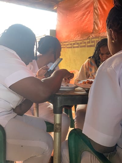 This image captures a group of four individuals seated around a rectangular dark-colored table with green plastic chairs, engaged in a meal or social gathering within a casual dining establishment in Akure, Nigeria. The setting appears to be a semi-outdoor area, indicated by bright daylight illuminating the scene and an overhead orange tarpaulin or fabric canopy.
Three of the individuals are dressed in white uniforms, consisting of white shirts and trousers, suggesting a professional or institutional affiliation. The person in the left foreground, with dark skin and braided hair, is actively holding and viewing a black smartphone, with their left arm resting on the table. Another individual in a white uniform is partially visible to the left, facing away from the camera. A third uniformed individual is seated in the right foreground with their back to the viewer.
The fourth individual, located in the right mid-ground, has dark skin, long braided hair, and is wearing a multi-colored patterned top. This person is looking down at a white plate containing food, which includes several fried, oblong pieces, likely plantains. Another white plate with food residue is visible on the table near the person with the smartphone. The background features a yellow wall adorned with a recurring brown geometric pattern, possibly a meander or Greek key design.