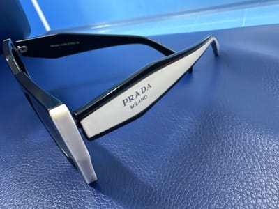 The image shows a close-up, angled view of a pair of black and white Prada sunglasses resting on a textured blue surface. The sunglasses are positioned with the temples folded in and are angled so that the right temple, which features "PRADA MILANO" in dark blue lettering on a white background, is most prominently displayed. The left temple is mostly obscured by the frame. The blue surface appears to be leather or a similar material with a distinct pebble-grain texture. In the background, a bright blue, possibly illuminated surface with a linear pattern is visible, suggesting a light source or a reflective surface. There are no people visible in the image. The scene is indoors, and the lighting is bright, creating reflections on the sunglasses. The image does not contain any information that would allow for its location to be identified as Orsk, Russia.