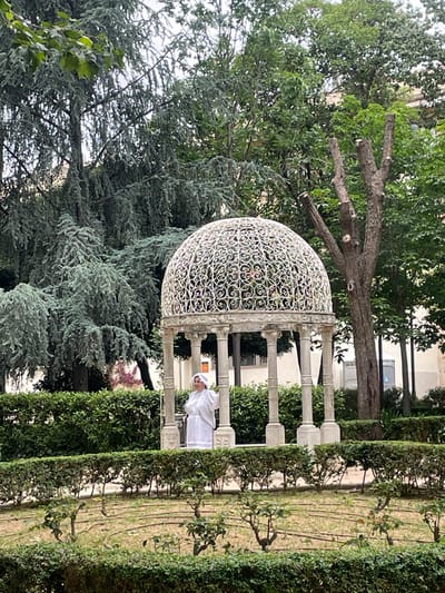 The image shows a garden scene in Terrassa, Spain. A white, ornate gazebo is surrounded by greenery, including tall hedges, trees, and bushes. A person dressed in white is partially visible within the gazebo. The garden setting includes pathways and trimmed vegetation. The trees have some branches that have been pruned. Buildings are visible in the background.