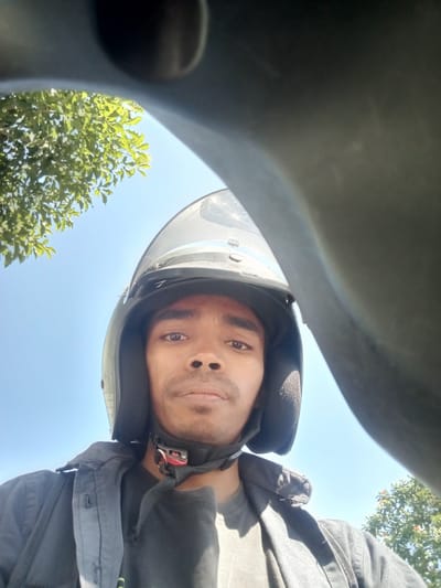 A medium shot from a first-person perspective shows a young man wearing a black helmet with a clear visor, partially obscuring his face. He is wearing a dark jacket over a dark shirt. The background consists of a bright blue sky with the leaves of a green tree visible on the left side. The image appears to have been taken outdoors, likely while on a motorbike, with a blurred dark object in the foreground suggesting a part of the vehicle. The overall lighting indicates it is daytime. The image does not contain any identifiable landmarks or contextual clues that would place it in Surabaya, Indonesia or any other specific location.