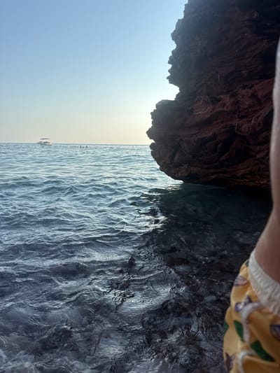 This media file depicts a person's legs in patterned shorts, with a rock formation and the sea in the background. The scene is shot from a low angle, looking downwards towards the water. The rock formation is dark and textured, appearing to overhang the water. The sea is calm with gentle ripples, reflecting the sunlight. In the distance, a small boat is visible on the horizon, suggesting the location is a coastal area. The sky transitions from a clear blue to a hazy yellow near the horizon, indicating it is likely daytime, possibly late afternoon or early morning. The overall mood is serene and relaxed, suggesting a leisurely moment by the sea. The orientation of the image is sideways. No text is visible.