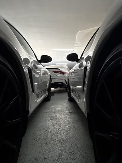 The image, rotated 90 degrees counter-clockwise, depicts a narrow indoor parking scene featuring two white cars. In the foreground, the right side and front of a white Porsche sports car are prominent, showcasing its glossy bodywork and a large black wheel. The text "PORSCHE" is clearly visible in dark, vertically oriented lettering on the car's front hood/frunk area. A black side mirror extends from the car's front-right fender.

Behind the Porsche, a second white car is parked, with its rear section and red taillights partially visible, indicating the extremely close proximity of the two vehicles. The setting is defined by contrasting walls: a smooth, light-colored wall on the left, reflecting ambient light, and a dark, textured concrete wall on the right. The floor is also dark and textured, consistent with a garage or parking facility. The lighting is artificial, suggesting an indoor environment. No specific activity is taking place beyond the cars being parked. The location is Cape Town, South Africa.