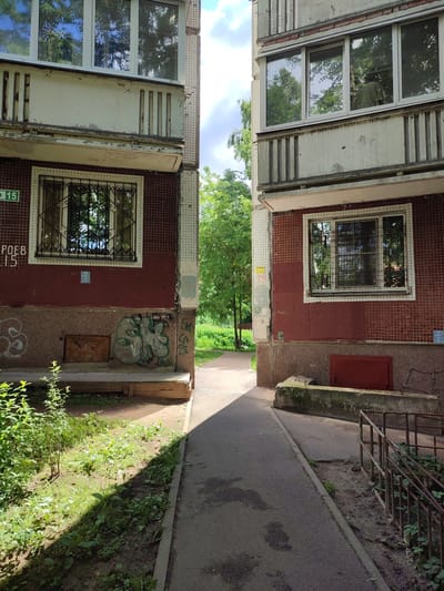 The image depicts the exterior of two adjacent apartment buildings in Vsevolozhsk, Russia, viewed from a pathway. The buildings appear to be constructed of concrete and tile, with some signs of wear and tear. Red tiles cover the lower portions of the buildings, while the upper sections are a lighter color, possibly painted concrete. Each building has several windows, some with metal grates. The pathway is asphalt, bordered by grassy areas and some overgrown vegetation. There is graffiti on the lower part of the left building. A metal railing is visible on the right side of the pathway, leading downwards. The sky above is partly cloudy with visible sunlight. The overall scene suggests a somewhat aged, urban residential environment.