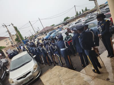A group of approximately 30 people, primarily men, are gathered outdoors, some in uniform and others in civilian attire. The individuals in uniform are wearing dark blue suits with light blue shirts and dark hats, some with gold epaulets and sashes. They appear to be in formation, standing at attention, and many are holding swords. In the foreground, a silver Toyota Yaris is parked, facing towards the right. Behind the uniformed individuals, a line of parked cars, including several blue sedans and silver vehicles, are visible. The environment suggests a street or an open area in front of a building. There are power lines and utility poles overhead, and trees are present in the background. The sky is overcast. The location is identified as the city of Barden Yana, Nigeria.