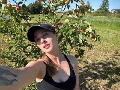 A woman wearing a black tank top and a black baseball cap takes a selfie in an orchard with apple trees laden with fruit. The woman is positioned in the foreground, slightly off-center, with her face partially obscured by the cap. Her eyes are blue, and her lips are slightly parted, revealing her teeth. She has a tattoo on her left arm, which is visible in the lower left corner of the image. The background consists of more apple trees, green grass, and a few distant structures and trees. The scene appears to be outdoors in a rural setting, consistent with the location of Турово (Лотошинский Район), Russia, known for its agricultural landscape. The lighting suggests it is daytime, with the sun casting shadows on the ground.