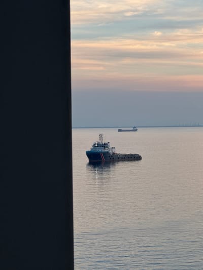 A tugboat is pictured in the foreground on a calm sea, with a larger cargo ship in the distance. The sky above is a soft blend of pastel colors, indicating either dawn or dusk. The scene is partially obscured by a dark, vertical structure on the left side of the frame. The location is identified as Unknown, Italy.