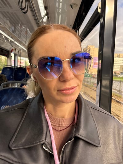 This is a portrait of a woman taken from a low angle, with the camera rotated. She is wearing blue mirrored sunglasses, a light pink ribbed turtleneck, and a dark leather jacket. The woman has blonde hair and is wearing a white earbud. She is looking towards the camera with her mouth slightly open, and her expression could be interpreted as relaxed or playful.
The background shows the interior of a vehicle, likely a train or bus, with rows of dark blue seats visible. Through the window, a cityscape with buildings is visible, suggesting an urban or suburban setting. The light indicates it is daytime. The overall impression is a casual, candid moment captured while traveling. There is no visible text in the image.