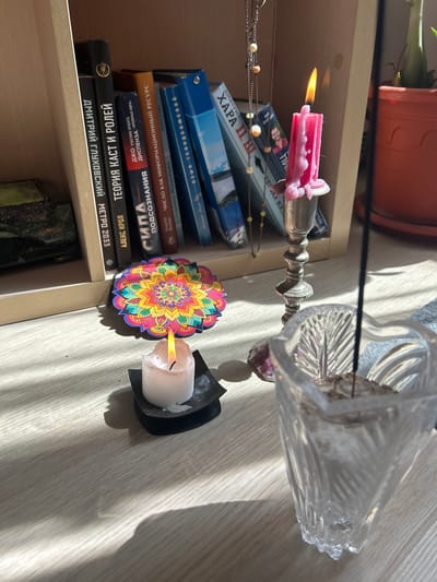 This media file captures a still life composition set on a wooden surface, likely a shelf or table. The scene is bathed in natural light, casting distinct shadows that suggest it's daytime, possibly late morning or early afternoon, with the sun filtering in through blinds or a window.

The primary objects are a stack of books, a vintage-style candlestick holding a lit pink candle, a second lit white candle in a dark holder, and a decorative mandala-like ornament. The books are in Russian, with titles such as "ДМИТРИЙ ГЛУХОВСКИЙ МЕТРО 2033" (Dmitry Glukhovsky Metro 2033), "ТЕОРИЯ КАСТ И РОЛЕЙ" (Theory of Cast and Roles), and "ЯЗЫК ТЕЛОДВИЖЕНИЙ" (Language of Body Movements). A delicate chain with beads is draped across some of the books. A clear glass vase, possibly containing dried flowers or an incense stick, sits to the right, with a pink crystal at its base.

The lit candles create a warm, ambient glow, contrasting with the cool light and shadows. The scene evokes a sense of quiet contemplation or a ritualistic setting, perhaps an altar or a personal space for reflection. The overall impression is one of a cozy, slightly mystical, indoor environment in Kemerovo, Russia.