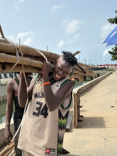 A man stands in Igboefon, Nigeria, carrying a bundle of bamboo. He wears a Milwaukee #34 basketball jersey, black shorts, and sandals. An orange bracelet adorns his right wrist. The bamboo is secured with white plastic strapping. A bridge, water, and a blue banner are visible in the background. The picture is taken in daylight.