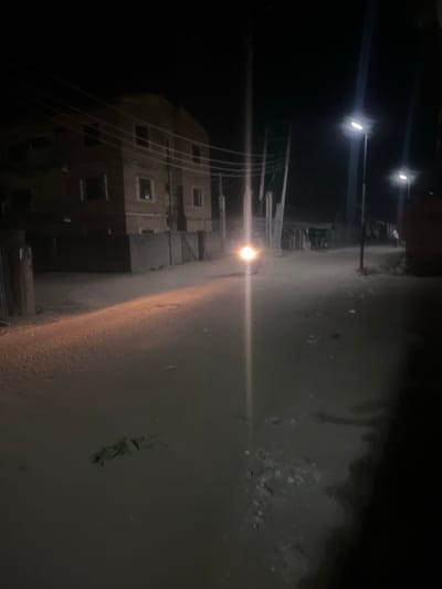 A dark, nighttime street scene reveals a building with lit windows on the left. Overhead power lines crisscross the sky. Streetlights illuminate the roadway, creating a bright glare. The road surface appears wet or slick, reflecting the light. A few small patches of vegetation are visible on the right edge of the street. The overall impression is of an urban or residential area at night.