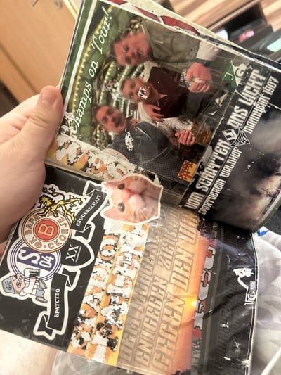 The image is a close-up, eye-level shot of a person holding an open scrapbook or magazine. The scrapbook is filled with various stickers and printed pages.
In the top half of the scrapbook, there is a printed image of three men, their faces pixelated, holding drinks. Text above this image reads "Champs on Tour!". To the right of this image, text reads "VOM SCHATTEN WALDHOF SPORTVEREIN WALDHOF MANNHEIM 1907".
The bottom half of the scrapbook features several stickers. One prominent sticker on the left shows a cartoon character holding a drink and wearing a hat. Next to it, there is a circular sticker with Cyrillic text and a "B" inside a circle, and a black banner with Cyrillic text that translates to "Brotherhood". Above these stickers, a sticker depicts a stylized yellow and black design. To the right of these stickers is a small sticker with an image of a cat's face. Further to the right, there are vertical strips with colorful, possibly abstract designs and more text.
The overall impression is that of a personal collection of memorabilia, possibly related to a sports club or a group of friends. The specific location in Gelsenkirchen, Germany, is not determinable from the visual information presented in the image.