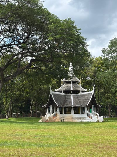 In Chiang Mai City Municipality, Thailand, an open grassy area is dominated by a traditional Thai pavilion with a multi-tiered, dark grey roof and white decorative trim. The pavilion is situated amidst lush green trees, with a large, sprawling tree with dark branches in the foreground on the left. The sky above is overcast with grey clouds. The pavilion has a white base adorned with white ornamental carvings resembling stylized birds or mythical creatures on either side of the entrance stairs. The surrounding environment is a park-like setting with well-maintained green grass. No people are visible in the scene.