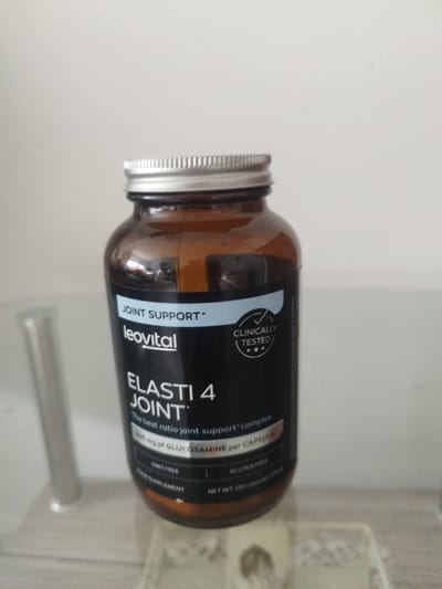 A brown glass bottle of Leovital Elasti 4 Joint supplement is placed on a reflective surface. The bottle has a silver metal cap and a black label with white text. The label reads "JOINT SUPPORT", "leovital", "ELASTI 4 JOINT", "The best ratio joint support complex", "500 mg of GLUCOSAMINE per CAPSULE", "GMO FREE", "GLUTEN FREE", "FOOD SUPPLEMENT", and "NET WT 120 capsules (120)". A portion of a silver metal object is visible to the left of the bottle. The background is a plain white wall. The scene appears to be indoors, possibly in a home or office setting. No specific location in Nuremberg, Germany is indicated by any visual cues in the image.