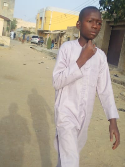 An outdoor scene in Garun Gawa, Nigeria, depicts a young male in the foreground walking on an unpaved, light brown dirt road. The individual, estimated to be 10-14 years old, wears a light purple or off-white long-sleeved tunic. His right hand is raised to his chin/neck area. His gaze is directed slightly to his right.
In the mid-ground, a silver SUV and a motorcycle are parked on the left side of the road. Several other individuals are visible in the distance: a group of adults in light-colored traditional attire, a woman in a dark green garment near a makeshift stall or structure, and a child near her. Two goats are also present on the dirt road.
The background features a mix of architecture. On the left, a light-colored building with a visible window is partially seen. Towards the center-right, a prominent two-story building with a bright yellow facade and a curved silver architectural element stands out. Other dilapidated structures with wooden frames and dark openings are scattered throughout the background. Utility lines are visible against the clear, light blue sky. Two distinct human shadows are cast on the dirt road to the left of the primary subject, indicating a high sun position.