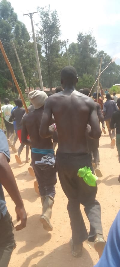 A group of people are walking on a dirt road. The men are all shirtless, with their backs facing the camera. They are walking in a line, with the man in the foreground walking ahead of the others. The men are wearing long pants and sandals or boots. The man in the foreground has a green towel hanging from his back pocket. The scene takes place in a rural area, with trees and power lines in the background. The road is dusty and brown. The location is Isukha ICHINA, Kenya, near coordinates 0.351, 34.779.