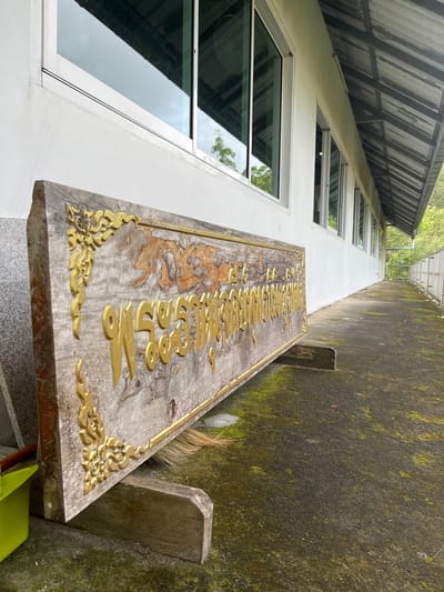 The image shows a weathered wooden sign with gold lettering and ornate gold corner decorations, resting on a wooden stand. The sign is positioned outdoors in front of a white building with a row of windows. The ground is a paved pathway with patches of moss. In the background, there is a railing and some greenery. The scene appears to be a walkway or entrance to a building, possibly a temple or a community center, given the style of the sign. The overall impression is one of a somewhat neglected but still functional outdoor area.