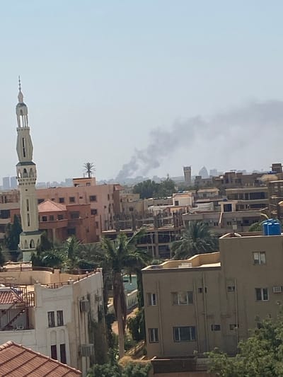 The image shows a rooftop view of the city of Omdurman, Sudan, at coordinates 15.683, 32.501. The scene is dominated by densely packed buildings, mostly multi-story apartment complexes, with a mixture of brick and concrete construction. The roofs of many buildings are visible, some with rooftop gardens or small structures. Palm trees and other vegetation are interspersed amongst the buildings, providing a touch of greenery. A tall minaret, likely belonging to a mosque, pierces the skyline in the foreground. Smoke billows in the distance, suggesting a possible fire or other activity. The sky is a pale blue, with a hazy quality, suggesting a dusty or smoky atmosphere. The overall impression is one of a bustling, densely populated urban environment. There are no people visible in the scene.