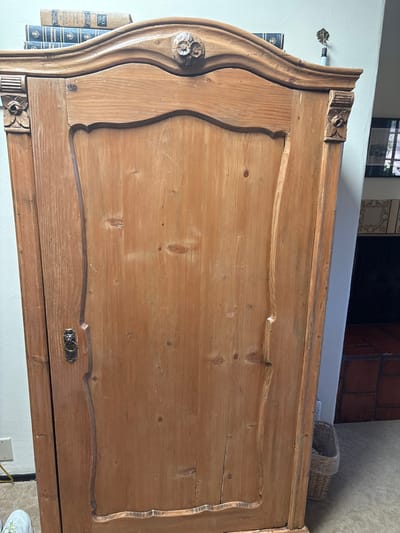 An antique-style wooden armoire stands against a light-colored wall in Monterey, United States of America. The armoire, constructed of light-toned wood (likely pine) with visible grain and knots, features a single door with a carved, undulating frame. The top is adorned with a curved pediment, incorporating a central carved medallion displaying a floral or stylized animal motif, flanked by carved details at the upper corners. A dark metal drop-ring handle is affixed to the left side of the door. The wood shows signs of wear, including scratches, minor indentations, and a vertical crack along the right edge of the door's carved frame. On top of the armoire, a stack of dark-bound books is visible to the left. To the right of the armoire, an adjacent area reveals reddish-brown terracotta floor tiles, patterned decorative wall tiles, and a dark recessed opening. A woven basket rests on a light-colored carpeted floor section in the lower right. In the lower left foreground, a portion of a light-colored shoe with green accents is visible, along with an electrical outlet from which a yellow cable extends. No individuals are depicted, and no specific actions or interactions are occurring.
