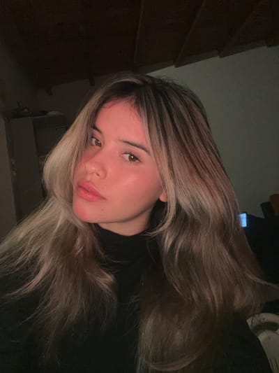 A young woman with blonde and brown ombre hair and fair skin poses for a selfie in an indoor setting. She is wearing a black turtleneck. Her hair is long and styled with layers. The lighting is dim, with some warm light illuminating her face. The background is dark and appears to be a room with wooden beams on the ceiling. No specific location in Sumalao, Argentina is identifiable.